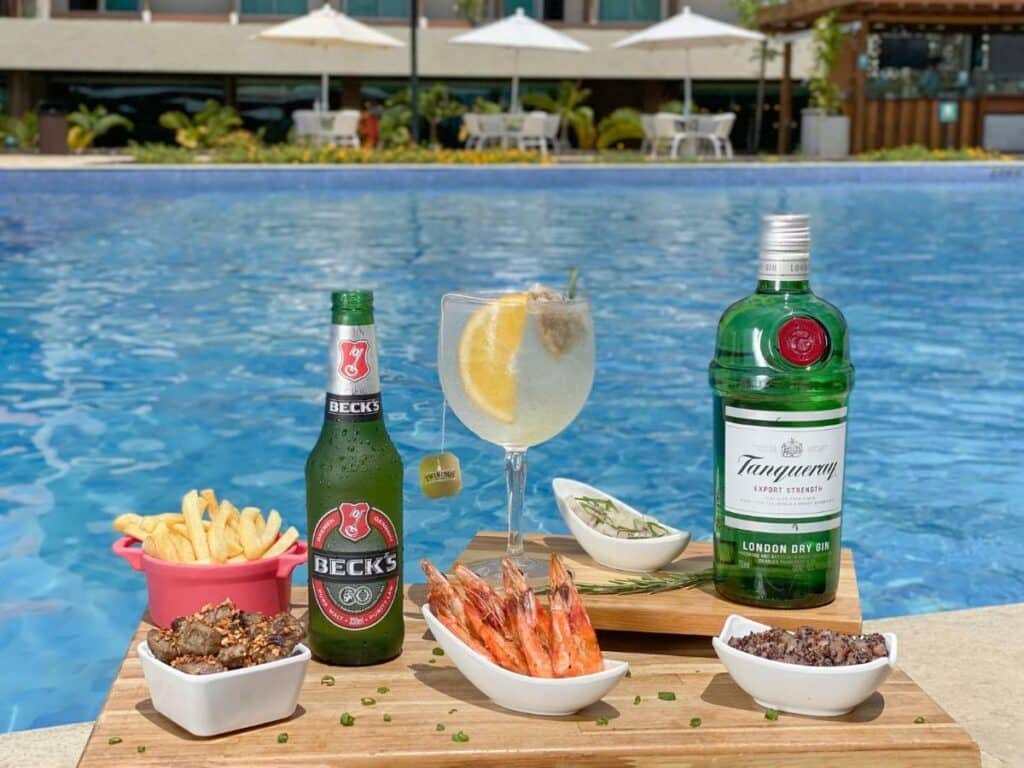 A imagem mostra uma mesa de madeira à beira da piscina, repleta de petiscos e bebidas, no Japaratinga Lounge Resort - All Inclusive, um dos melhores resorts all inclusive no Brasil. Em destaque, há uma garrafa de cerveja Beck