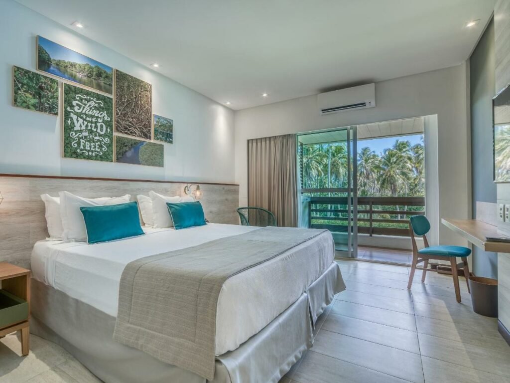 A imagem mostra um quarto do Japaratinga Lounge Resort - All Inclusive, um dos melhores resorts all inclusive no Brasil. Há uma cama de casal grande, com roupa de cama branca e travesseiros com capas azuis. Acima da cama, há uma composição de quadros com imagens da natureza e uma frase decorativa. À direita, há uma porta de vidro que dá acesso a uma varanda com vista para uma área arborizada com muitas palmeiras. O quarto também possui uma mesa de trabalho com cadeira e um ar-condicionado na parede, proporcionando um ambiente confortável e acolhedor.