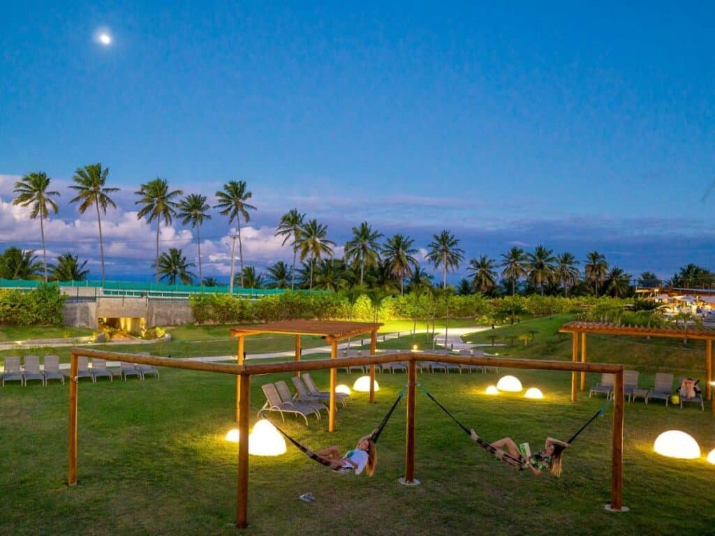 A imagem mostra uma área externa do Japaratinga Lounge Resort - All Inclusive ao entardecer. No gramado, há várias redes penduradas em estruturas de madeira, onde algumas pessoas relaxam. Ao fundo, palmeiras altas são visíveis, alinhadas em frente a um céu azul escuro com a lua brilhando. Há várias espreguiçadeiras espalhadas pelo gramado e iluminação suave vinda de lâmpadas no chão, criando um ambiente tranquilo e relaxante. Ao longe, é possível ver parte da infraestrutura do resort e a vegetação ao redor, com a paisagem do mar no horizonte.