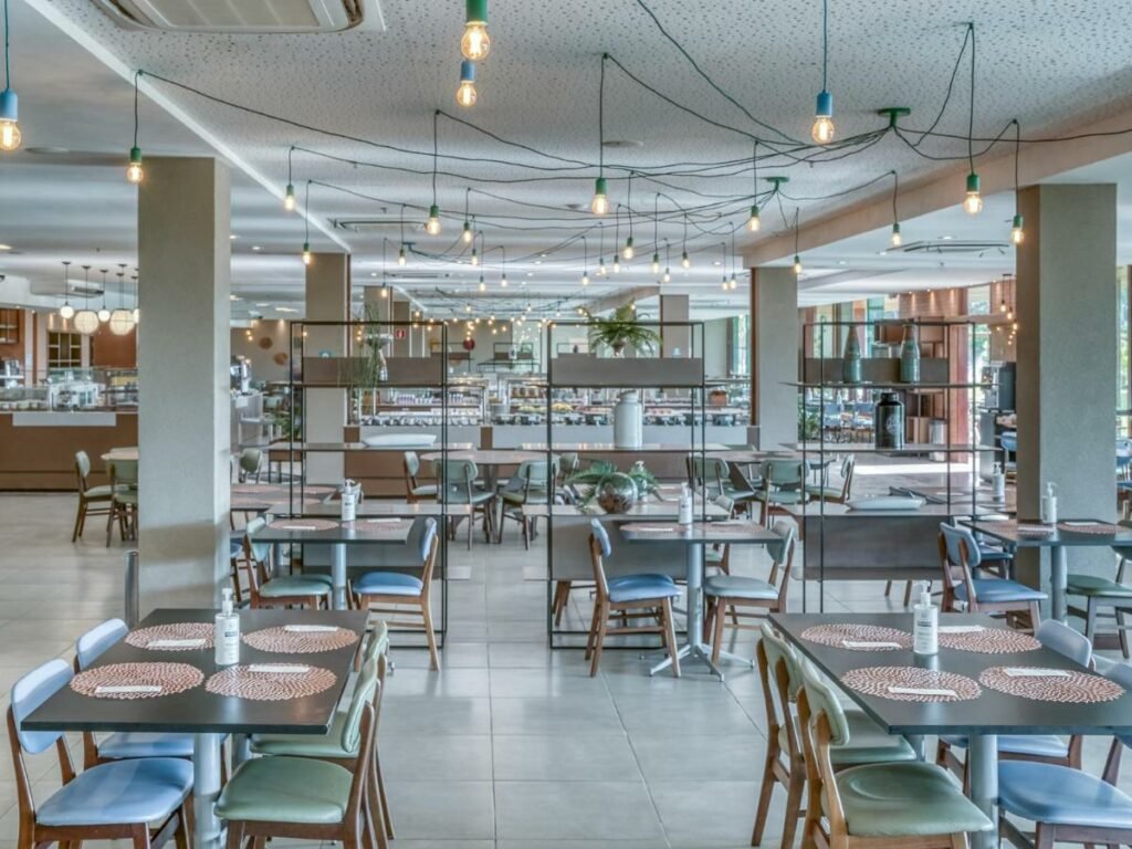 A imagem mostra o interior de um restaurante espaçoso e moderno no Japaratinga Lounge Resort - All Inclusive, um dos melhores resorts all inclusive no Brasil. As mesas, que acomodam de quatro a seis pessoas, estão organizadas de forma a proporcionar um ambiente aberto e confortável. Cada mesa possui jogos americanos decorativos e frascos de álcool em gel. O teto é adornado com várias lâmpadas penduradas, criando uma iluminação agradável e acolhedora. Ao fundo, é possível ver a área de buffet com uma grande variedade de alimentos dispostos. O ambiente é decorado com prateleiras contendo plantas e objetos decorativos, conferindo um toque de frescor ao local.