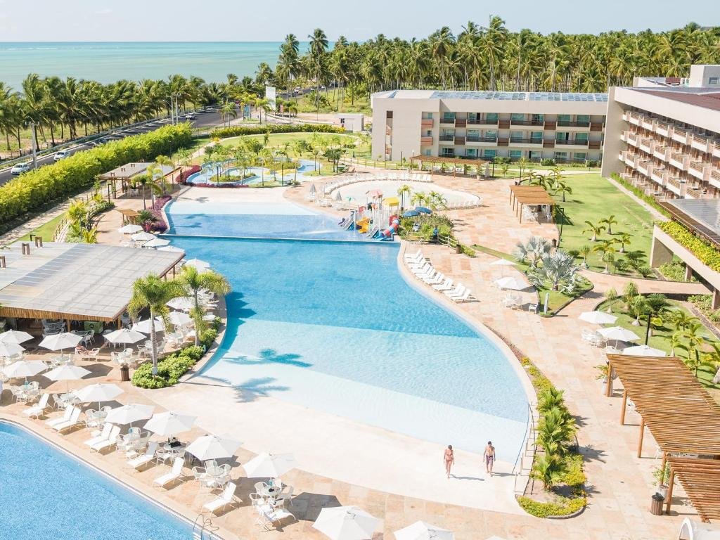 Área externa do Lounge Resort, com piscina extensa, cadeiras, espreguiçadeiras e guarda-sóis em volta, além de alguns jardins e o prédio do hotel no lado direito. No lado esquerdo há uma avenida separando o hotel de uma praia cristalina de água azulada