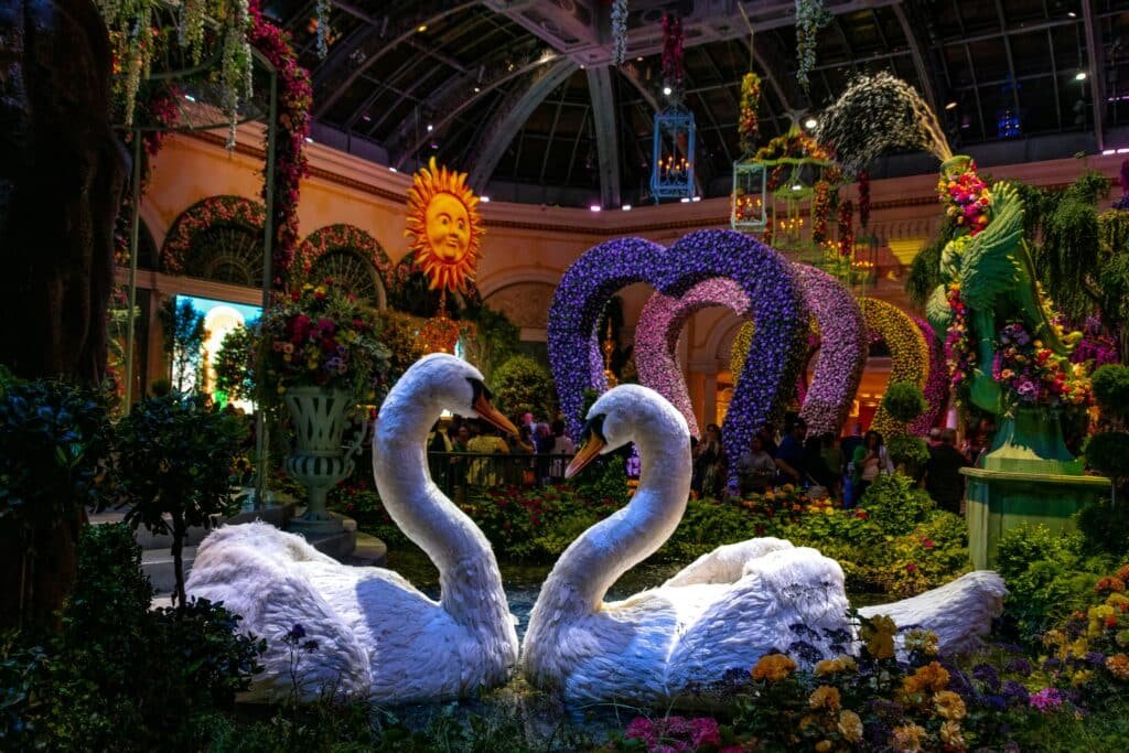 A foto mostra o jardim interno do Bellagio durante a noite. Em primeiro plano estão dois cines, um de frente para o outro, no meio de várias plantes e flores. Ao fundo estão vários coraçãoes de flores formando um túnel. O local é todo decorado com flores no chão e penduradas no teto, que é de vidro.