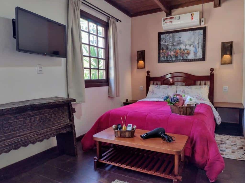 Quarto no Jardins de Canela - Aptos e Casas. A cama de casal está no centro da foto, e possui uma cesta com amenidades em cima. Na sua frente, há um banco com um secador de cabelo e outra cesta com utilidades. à sua esquerda há uma TV na parede e uma janela. Representa o post sobre airbnb em Canela.