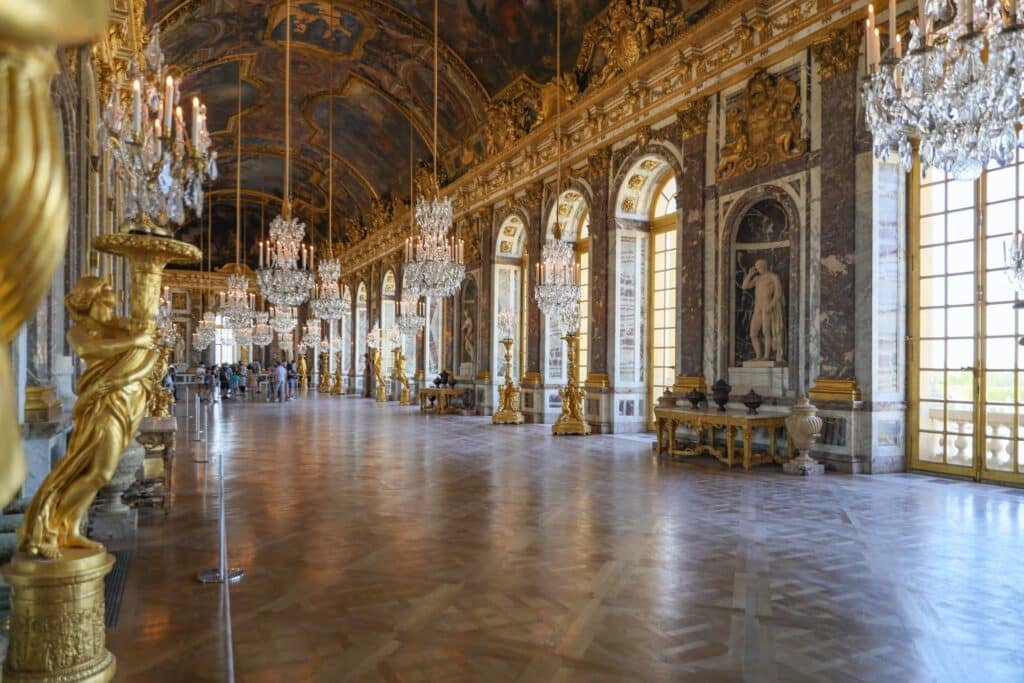 Lustres de cristal com velas pendurados na Sala dos Espelhos, no Palácio de Versalhes, uma das opções de o que fazer em Paris. As paredes decoradas com painéis possuem janelas e espelhos, além de estátuas e outras obras de arte. O teto abobadado também possui pinturas.