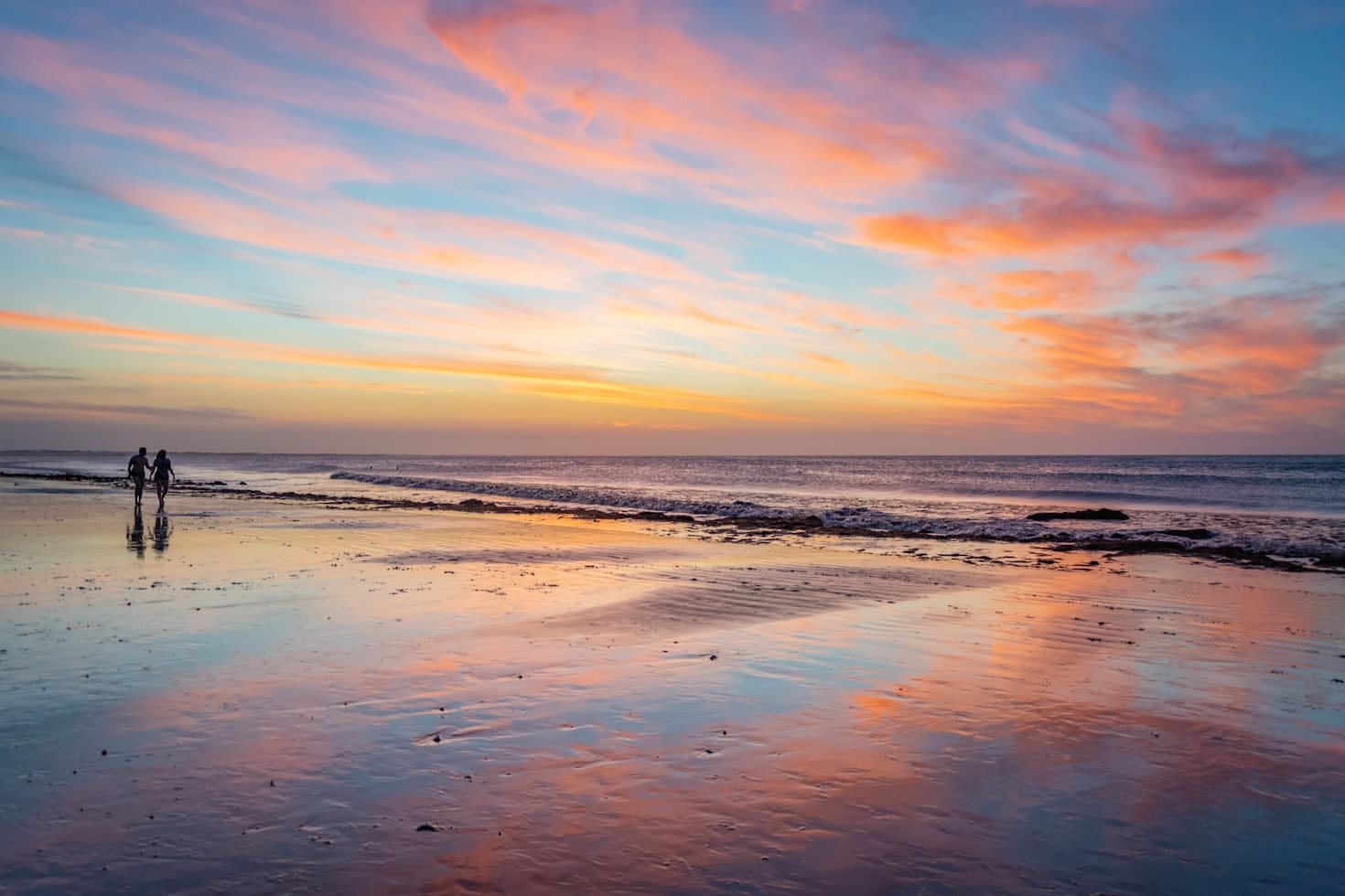 Um casal caminha pela praia ao pôr do sol, com nuvens coloridas refletindo na areia molhada e no mar calmo para representar um dos melhores lugares para lua de mel