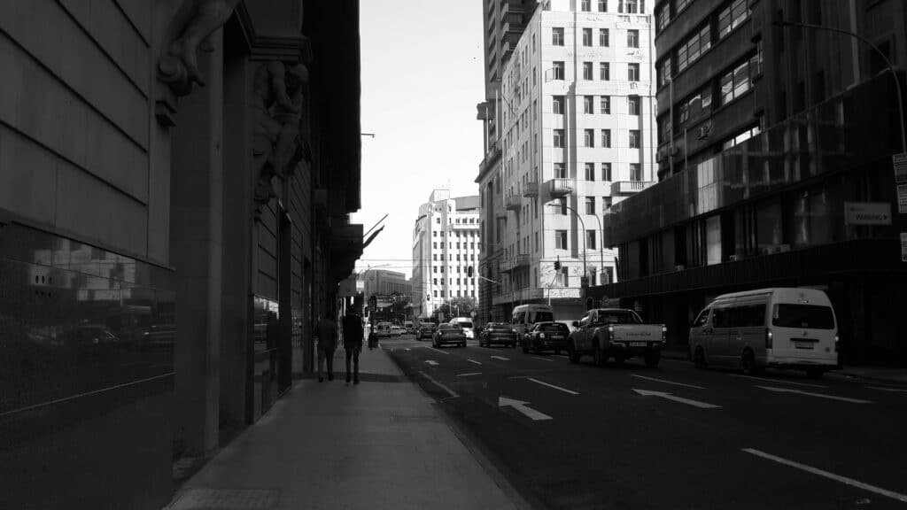 Imagem em preto e branco de uma rua urbana com carros, uma van e pessoas caminhando na calçada ao lado de edifícios altos em Joanesburgo.