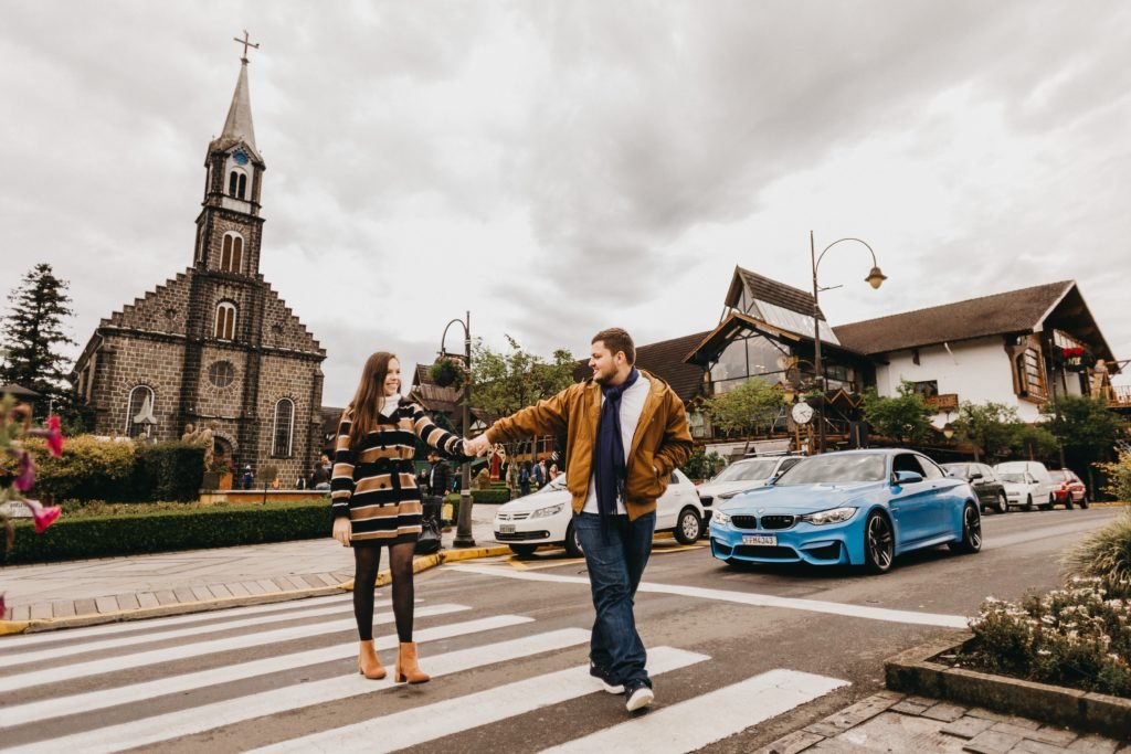 Um homem e uma mulher de mãos dadas enquanto atravessam uma faixa de pedestres em frente a paróquia de são pedro em gramado, por perto há carros estacionados, sob um céu nublado.