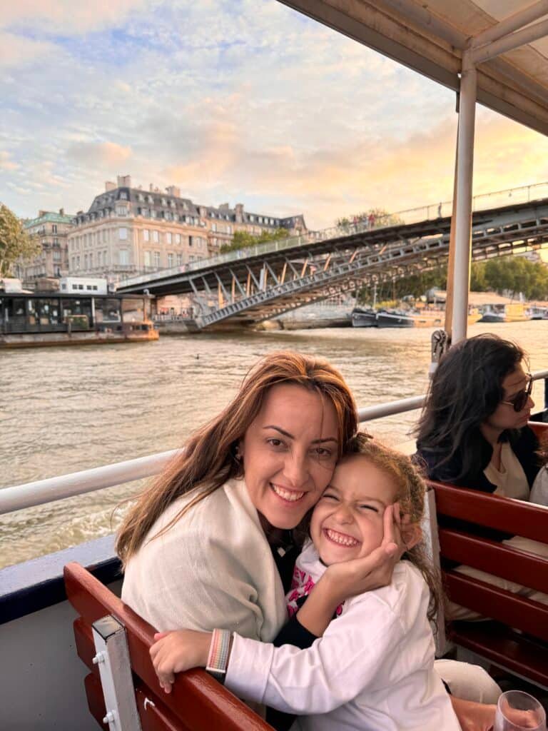 Uma mulher e uma criança sorriem e se abraçam em um barco, com edifícios e uma ponte sobre um rio visíveis ao fundo.