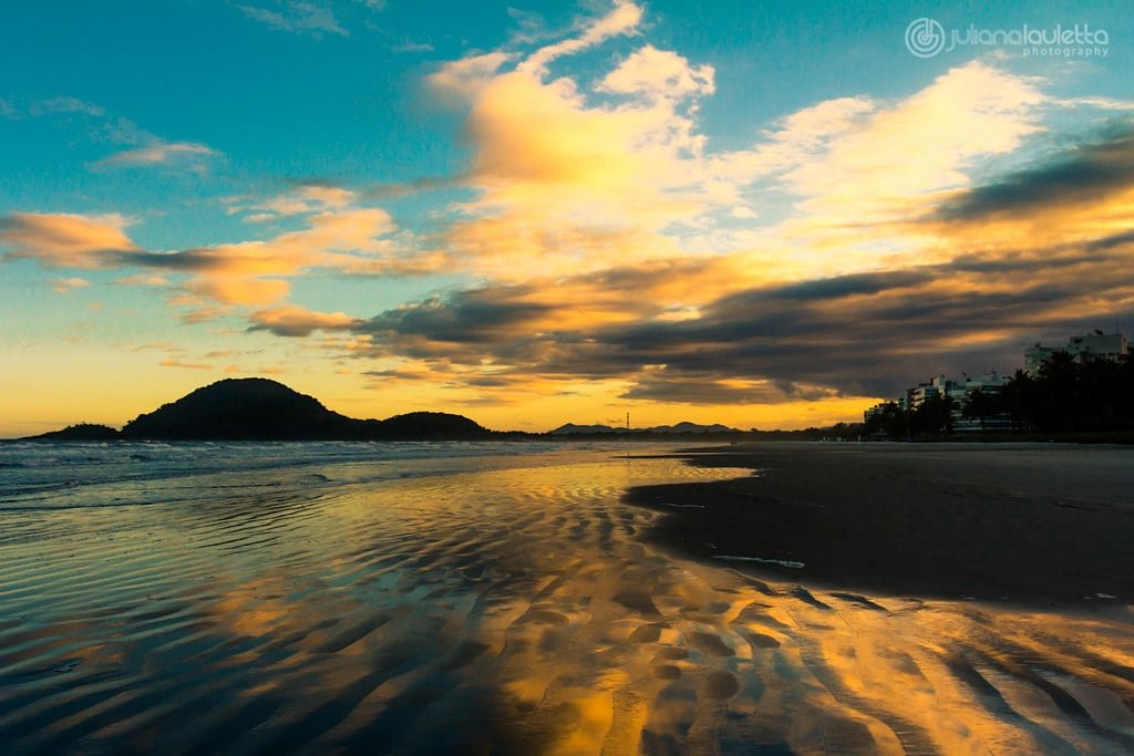 Pôr do sol sobre uma praia com areia ondulada em primeiro plano, água à esquerda e colinas recortadas sob um céu nublado.
