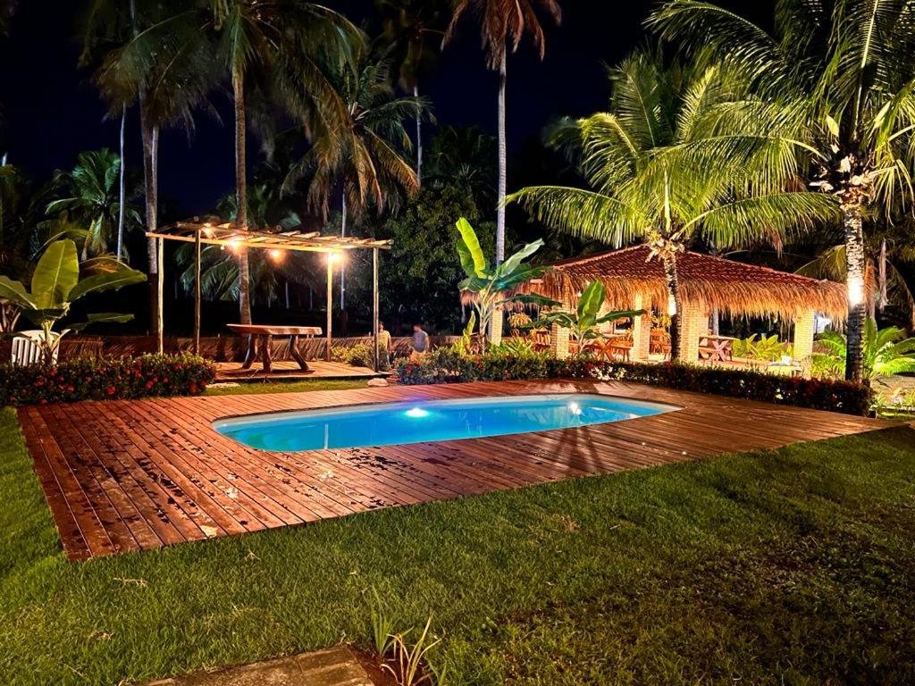 Piscina iluminada na área externa da pousada Kaçuá Milagres, com deck de madeira no mesmo nível do gramado, pergolado com mesa e alguns coqueiros próximos, já à noite