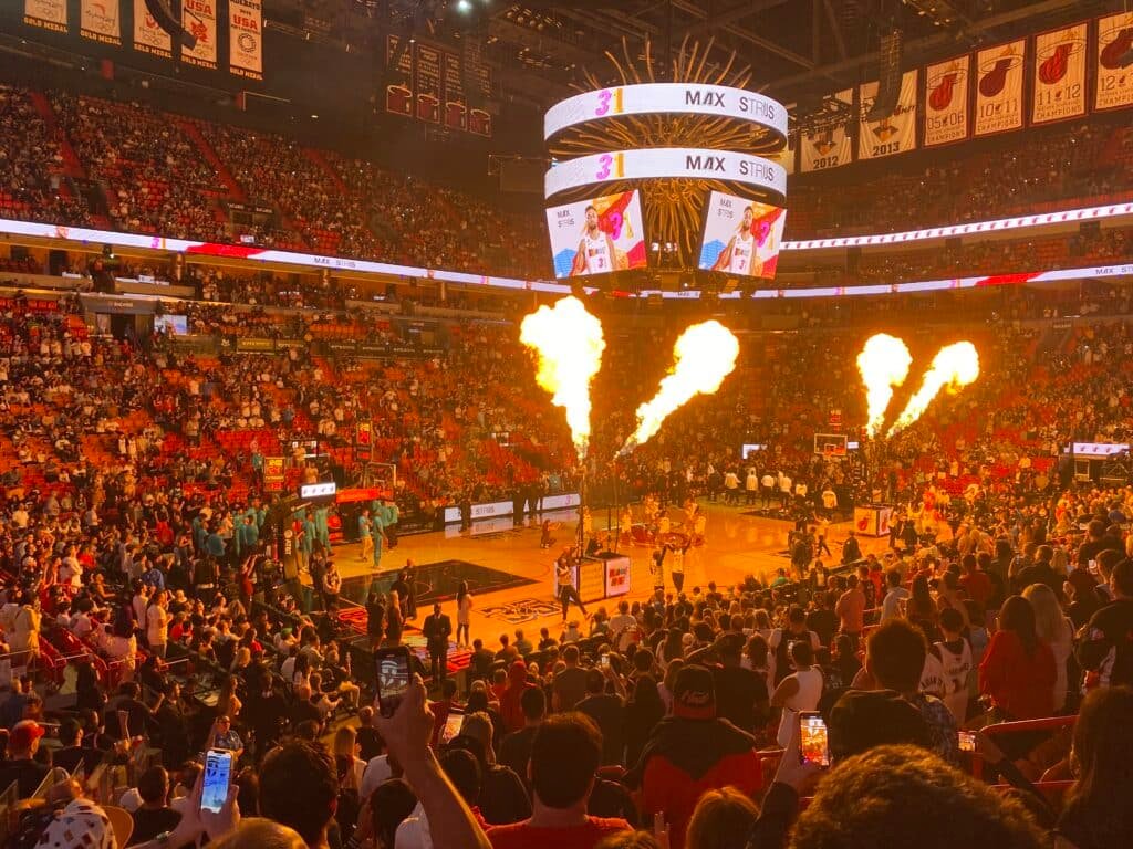 Arena Kaseya Center durante a apresentação de jogadores e abertura de um dos jogos do Miami Heat. As arquibancadas estão quase cheias e prestando atenção nas apresentações com fogo e líderes de torcida no centro. No telão acima passa a imagem de um jogador. A foto mostraa mais uma opção de o que fazer em Miami.