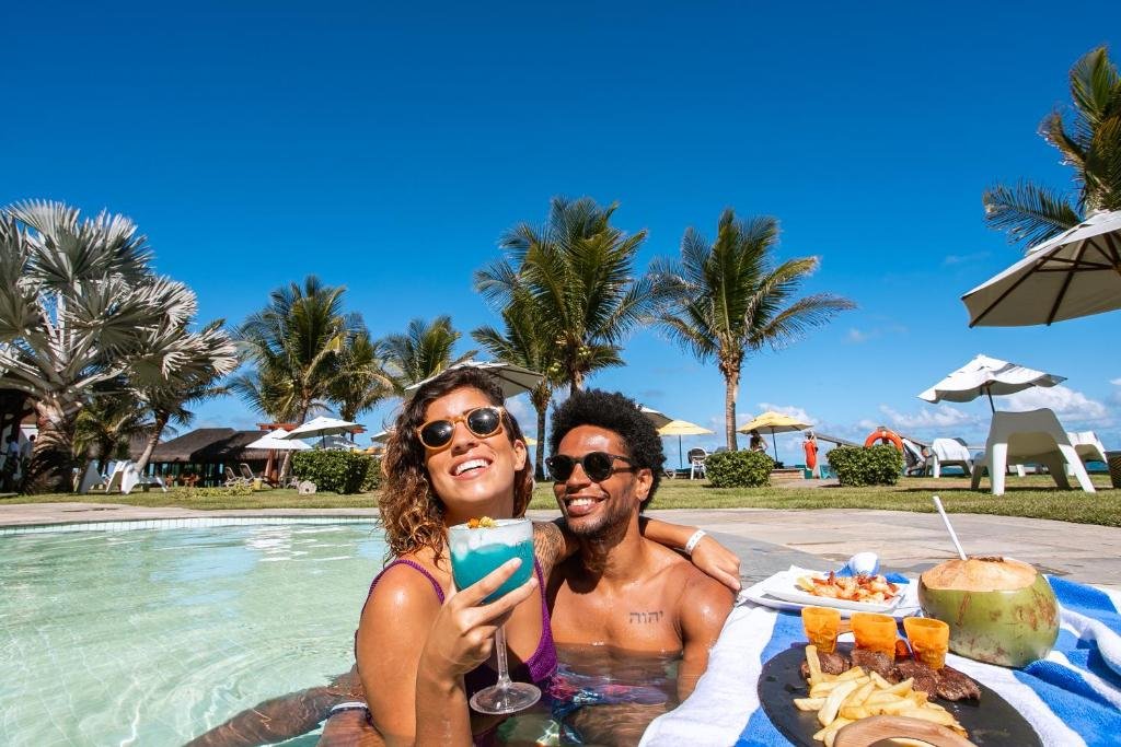 Um casal sorridente desfruta de drinques em uma piscina em um dia ensolarado. Palmeiras e espreguiçadeiras com guarda-sóis são visíveis ao fundo.