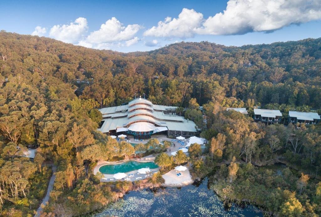 Vista aérea do Kingfisher Bay Resort cercado por uma densa floresta, com um edifício principal curvo, uma piscina e edifícios menores espalhados.