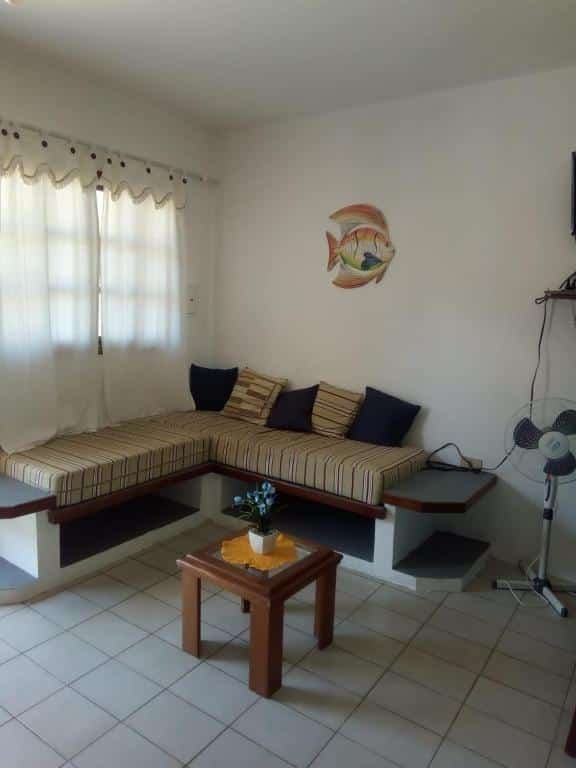 Sala de estar do kitnet Raquel. No centro uma mesinha, no fundo um sofá cama, no lado direito um ventilador e do lado esquerdo uma janela.