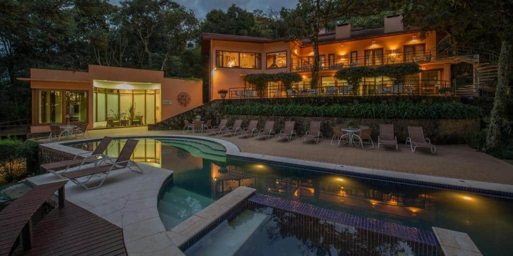 Pátio do Kuriuwa Hotel em Monte Verde com grandes janelas e varandas, cercada por árvores, com piscina com espreguiçadeiras e iluminação externa ao anoitecer.