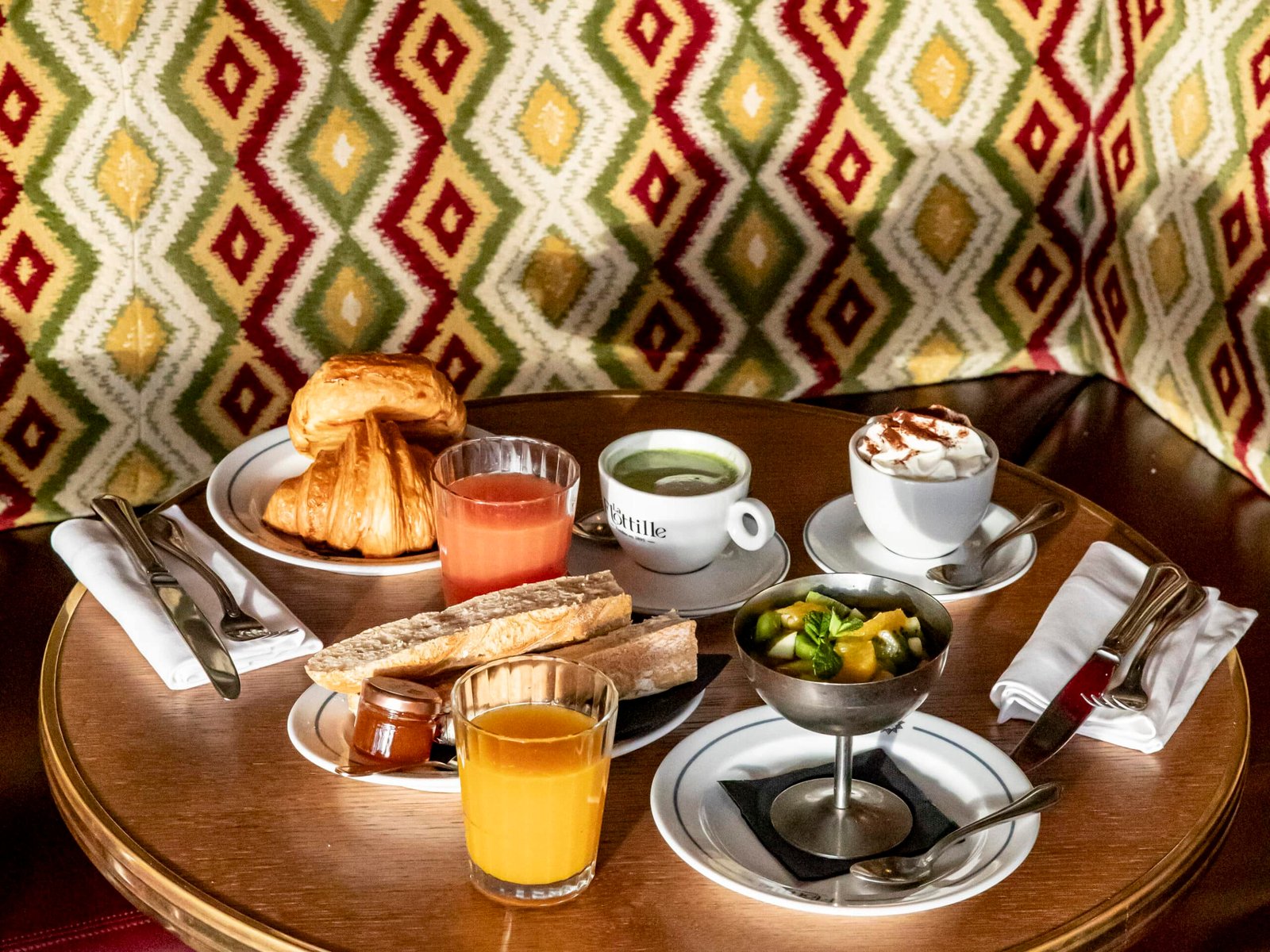 Uma pequena mesa redonda no restaurante La Flotille em Versalhes com vários itens de café da manhã, incluindo doces, bebidas e frutas, em frente a um estande estofado estampado.