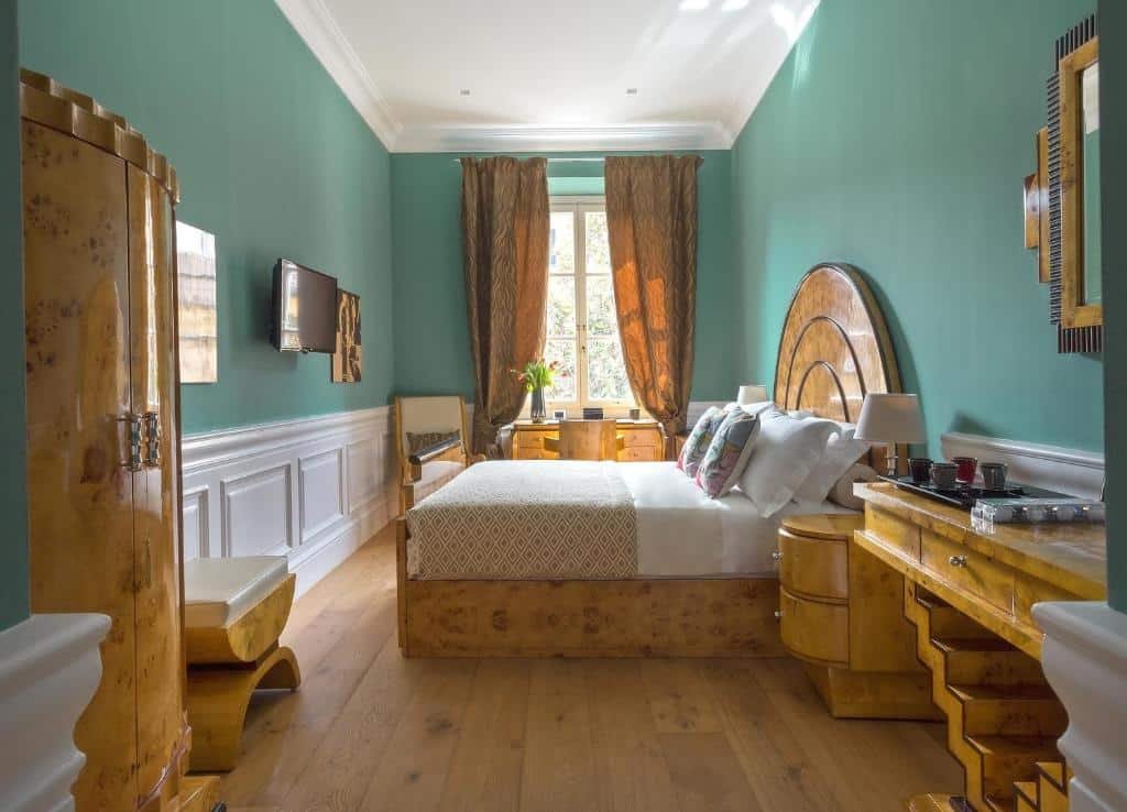 Quarto superior com cama queen-size do La Maison du Sage, com móveis de madeira clara, mesinha com xícaras em cima, cadeira, TV e mesa com cadeira em frente de uma janela iluminando o quarto com o raio do sol