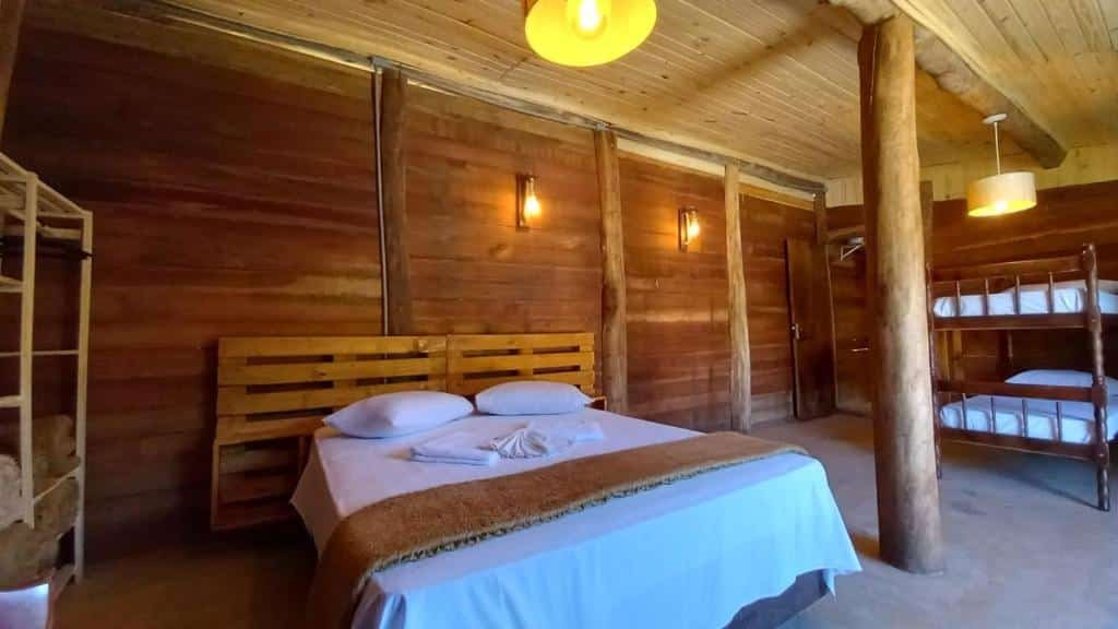 Foto do quarto em La Villa Chalés Garden. O chalé tem paredes de madeira. Há uma cama de casal no meio, uma estante à sua esquerda e na direita há uma beliche.
Ilustra o post sobre airbnb em Serra Negra.