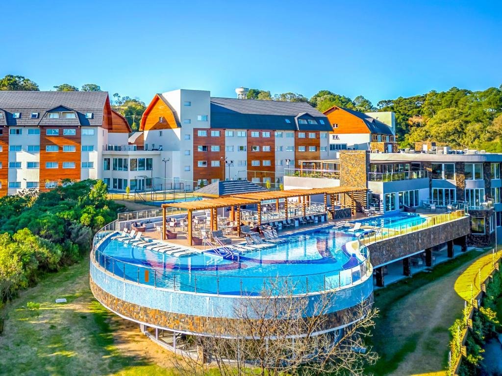 Um moderno complexo hoteleiro com piscina externa curva, cercado por vegetação e prédios de vários andares sob um céu azul claro.