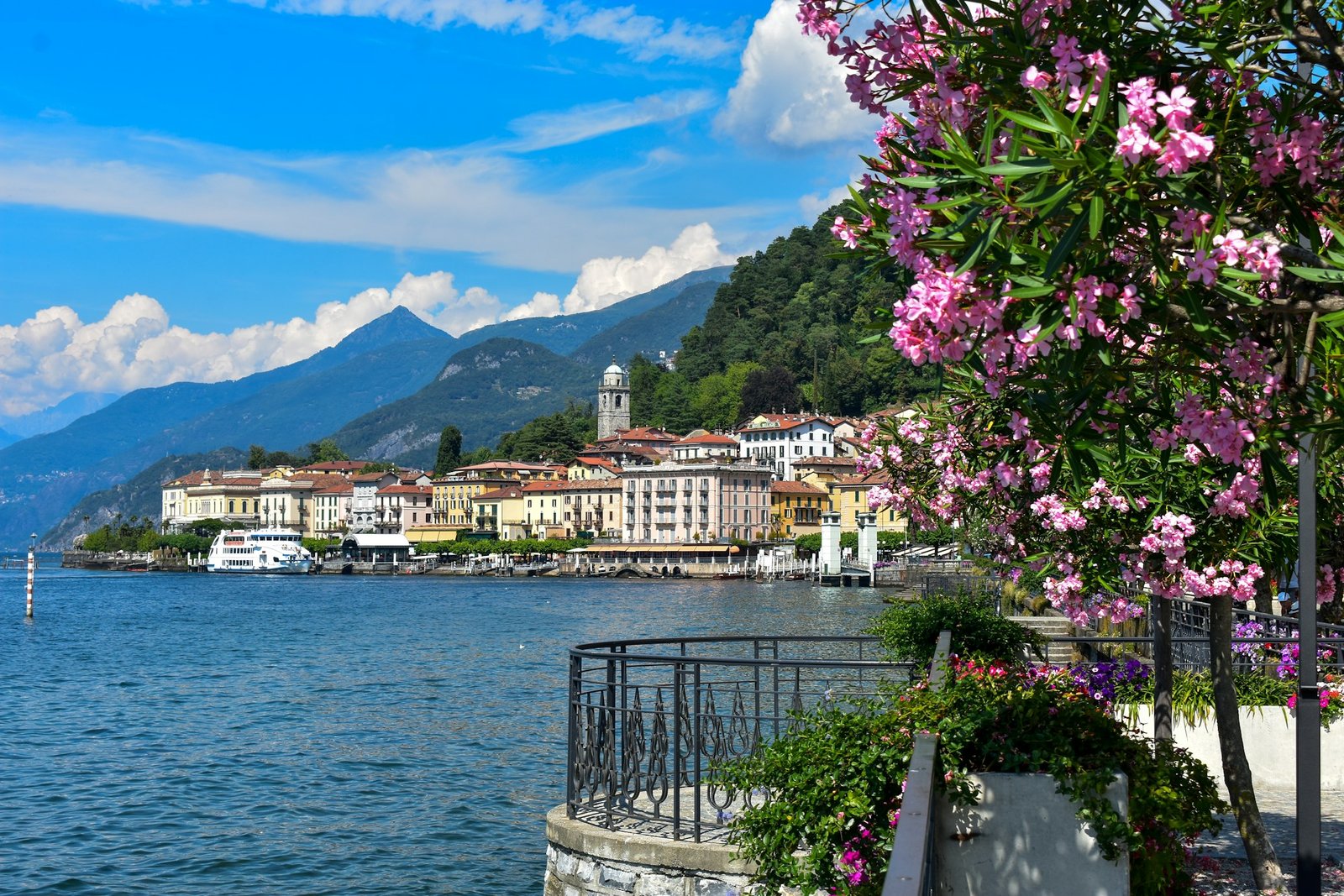 Lago di Como com edifícios coloridos e montanhas ao fundo, emolduradas por flores e uma grade em primeiro plano.