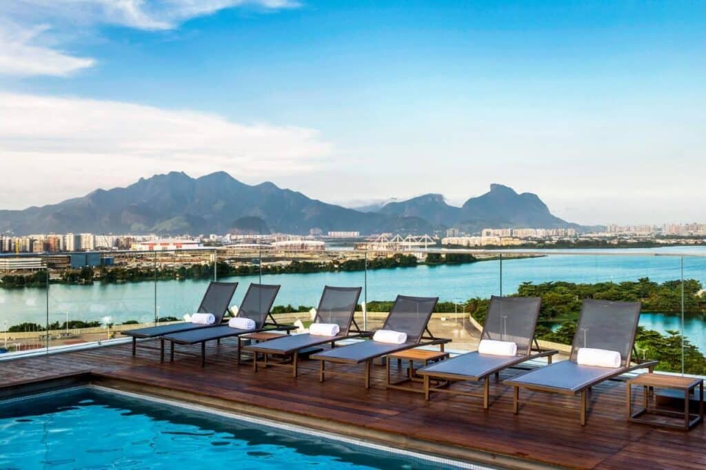 Área de piscina na cobertura do Lagune Barra Hotel com várias espreguiçadeiras com vista para o horizonte da cidade, montanhas e um grande corpo d