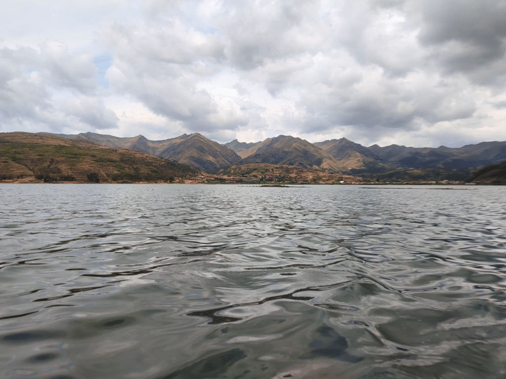 vista panorâmica do lago Piuray no Vale Sagrado dos Incas. A parte debaixo da imagem está tomada por águas escuras, enquanto da metade pra cima é possível ver várias montanhas e um céu nublado, carregado de de nuvens