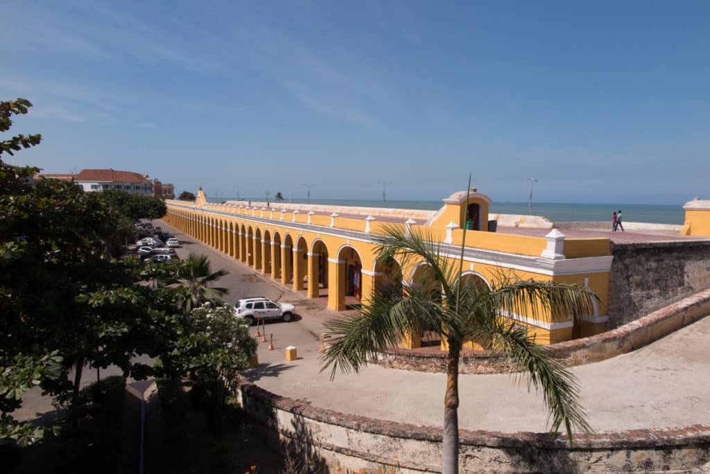 A imagem mostra um longo edifício colonial amarelo em Las Bóvedas com uma série de arcos que formam uma arcada. O edifício está localizado próximo ao mar, visível ao fundo, e é cercado por uma estrada onde alguns carros estão estacionados. Há vegetação verde e palmeiras ao redor, adicionando um toque tropical à cena. O céu é claro e azul, sugerindo um dia ensolarado.