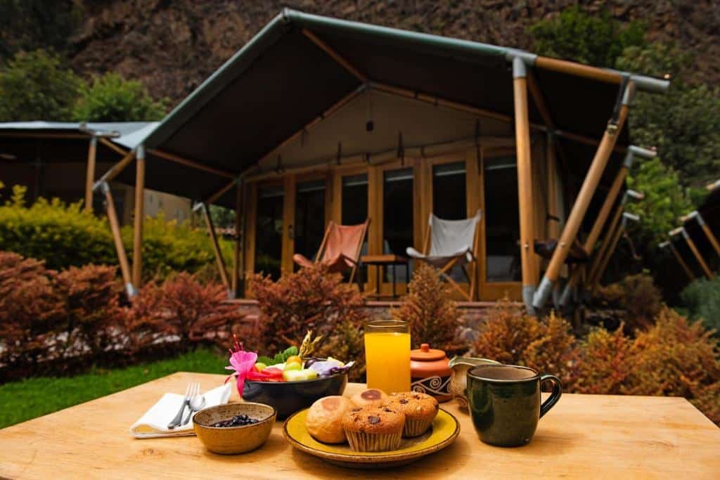 mesa de café da manhã ao ar livre do Las Qolqas EcoResort Ollantaytambo, com um prato de muffins, copo de suco de laranja e xícaras rústicas ao lado. Atrás da mesa é possível ver a tenda dos quartos em formato de chalé.