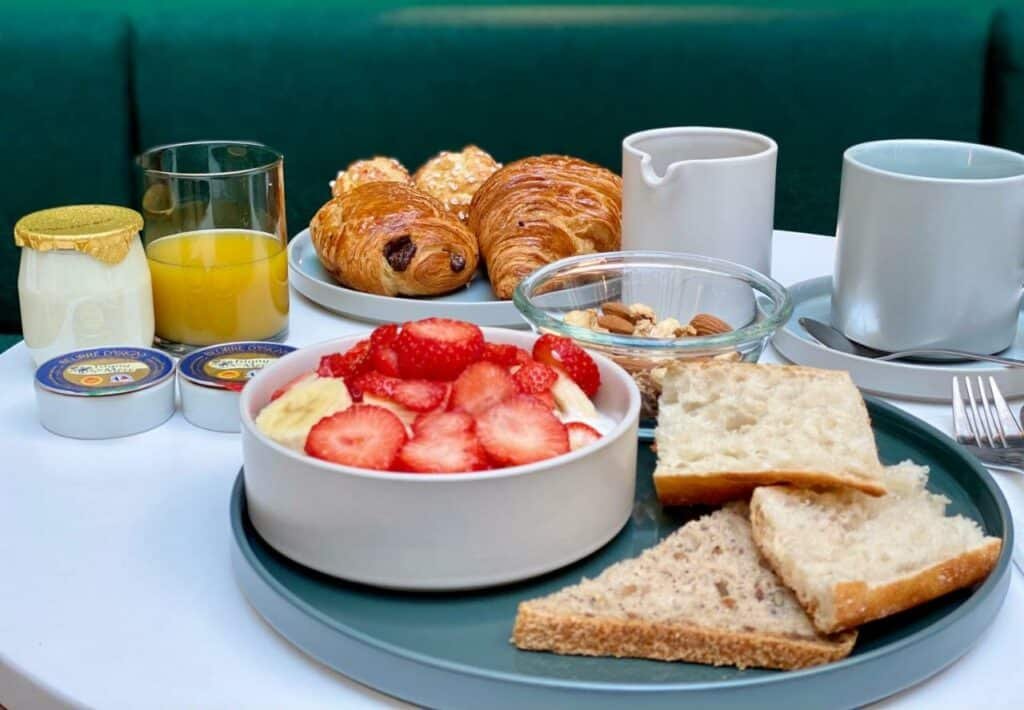 A imagem mostra uma mesa de café da manhã com uma variedade de itens no Le Basile Hôtel. Há um prato com pedaços de pão e torradas, uma tigela de iogurte com fatias de morango e banana, um copo de suco de laranja, e pequenos potes de geleia e manteiga. Ao fundo, há um prato com croissants e pães de chocolate. Também estão presentes uma tigela de nozes e amêndoas, uma caneca branca, uma jarra de leite e um pote de iogurte. A mesa tem uma aparência organizada e apetitosa, criando um ambiente convidativo para a refeição.