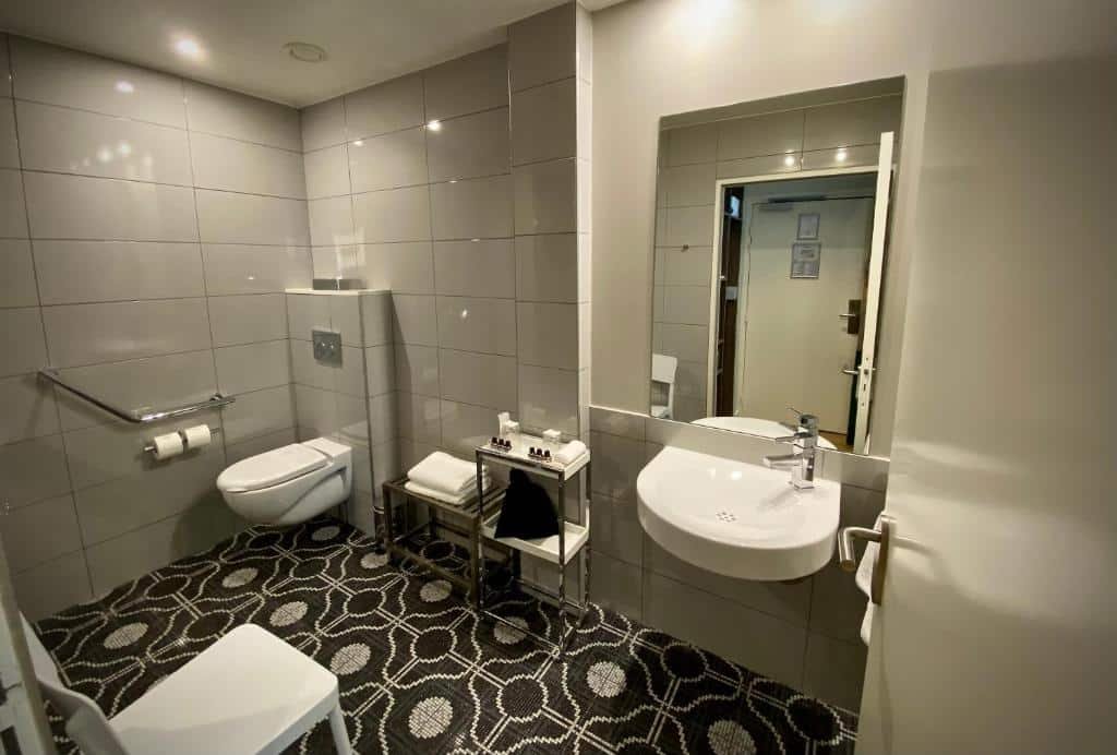 A imagem mostra um banheiro adaptado para pessoas com deficiência no Le Basile Hôtel. As paredes são revestidas de azulejos cinza claro, e o chão tem um padrão geométrico preto e branco. Há um vaso sanitário com barras de apoio ao lado, e ao lado dele, um suporte com toalhas e produtos de higiene. A pia é suspensa, permitindo acesso fácil, e acima dela há um grande espelho. Há também uma cadeira branca ao lado da pia. O banheiro é espaçoso e bem iluminado, projetado para ser acessível e funcional.