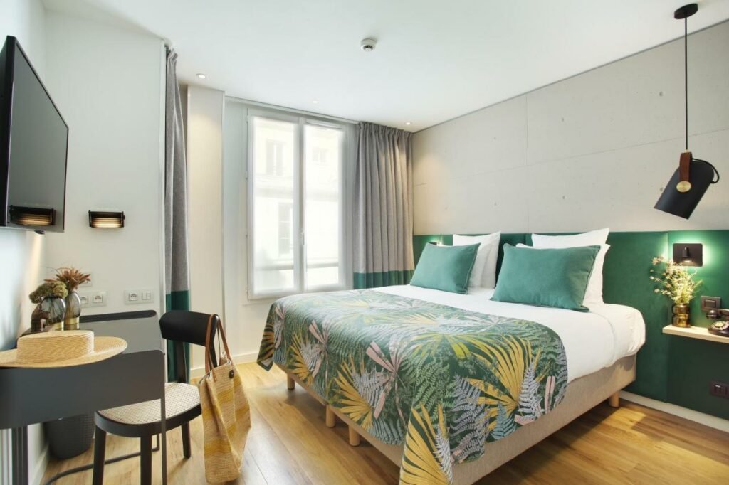 A imagem mostra um quarto moderno e bem iluminado no Le Basile Hôtel, um dos hotéis com café da manhã em Paris. A cama é grande, com roupa de cama branca e uma colcha de padrão tropical verde e rosa, combinando com as almofadas verdes. Há uma janela alta com cortinas cinza que permitem a entrada de luz natural. Ao lado da cama, há uma prateleira com uma lâmpada moderna e um pequeno vaso com flores. Uma mesa pequena com cadeira preta e um chapéu de palha complementam a decoração minimalista. O chão é de madeira clara, criando um ambiente acolhedor e relaxante.