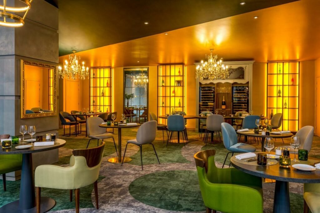 Restaurante do Le Louis Versailles Château com mesas e cadeiras dispostas ao redor. Alguns lustres de vidro estão pendurados acima das mesas. Na parede ao fundo há prateleiras com garrafas de vinho e um armário de madeira. Já numa coluna ao lado esquerdo fica um espelho quadrado.