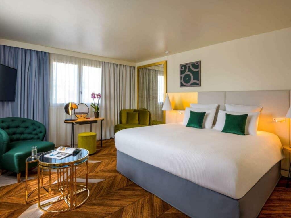 Quarto do Le Louis Versailles Château, um dos hotéis perto do Palácio de Versalhes. Uma cama de casal com mesinhas de cabeceira e abajures dos dois lados está encostada na parede ao lado direito. Em frente está uma mesinha redonda e uma poltrona com uma TV logo acima. Ao lado da poltrona há uma pequena penteadeira com espelho, banquinho e vaso de flores, e uma janela com cortinas fica logo atrás dessa mesa. Na mesma parede da cama há um grande espelho, e em frente a ele há uma poltrona com almofadas.