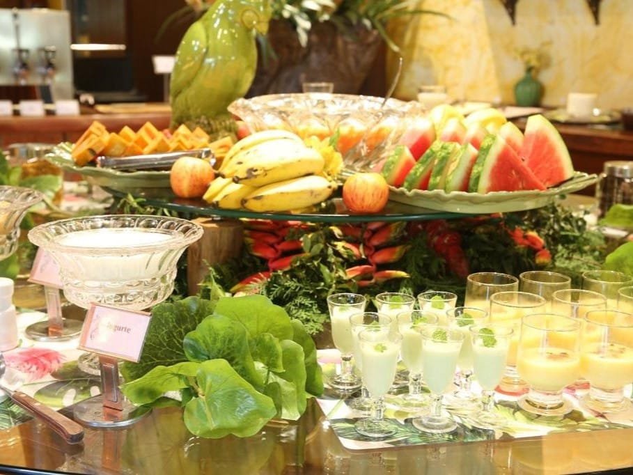 A imagem mostra uma mesa de buffet rica em frutas e sobremesas frescas. No centro, há uma bandeja com bananas, maçãs e fatias de melancia e mamão. Ao redor, copos de vidro contêm bebidas ou sobremesas cremosas, decoradas com folhas de hortelã. Há também uma tigela de iogurte ao lado de folhas verdes, possivelmente usadas para saladas. A apresentação é exuberante, com uma decoração tropical que inclui um grande papagaio de cerâmica verde no fundo. Tudo está organizado de forma colorida e atrativa. Esse é o Leão de Judá Charme Hotel, um dos hotéis-fazenda em Minas Gerais.