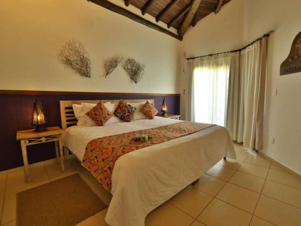 A imagem mostra um quarto aconchegante com decoração rústica e detalhes coloridos no Leão de Judá Charme Hotel. No centro, há uma cama de casal ampla, coberta com uma colcha branca e adornada com almofadas em tons vibrantes e um trilho de cama com estampa étnica. As paredes são brancas, e no alto da cama, há três enfeites decorativos que lembram galhos secos. O teto de madeira tem vigas aparentes, e o ambiente é iluminado por abajures rústicos sobre mesas laterais. Cortinas claras cobrem uma janela grande que deixa a luz natural entrar no quarto.