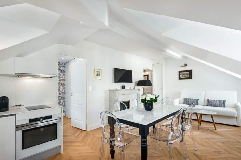 Interior do Les Demoiselles à Versailles. Na lateral esquerda é possível ver parte da cozinha, onde fica o fogão e uma cafeteira. No centro da foto está uma mesa de jantar com seis cadeiras, e ao fundo, na sala de estar, um sofá com almofadas está ao lado de uma lareira que tem TV acima e abajur ao lado.