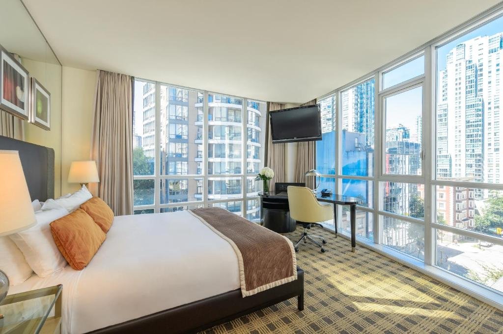 Quarto no Loden Vancouver com duas paredes feitas de vidro com janelas dando vista para a cidade, há cortinas para fechar essa parte de vidro, o chão é de carpete com desenho xadrez, há uma cama de casal com almofadas e travesseiros, no canto do quarto é possível ver uma pequena mesa de escritório e uma cadeira giratória, sob essa mesa presa na parede, há uma televisão, para representar hotéis em Vancouver