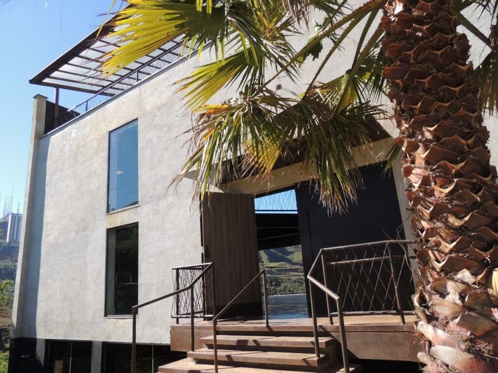 Frente do edifício do Loft Espaço Vila da Serra, com uma árvore do lado direito e uma pequena escada dando acesso ao prédio que é alto, estilo industrial, e tem uma cobertura coberta