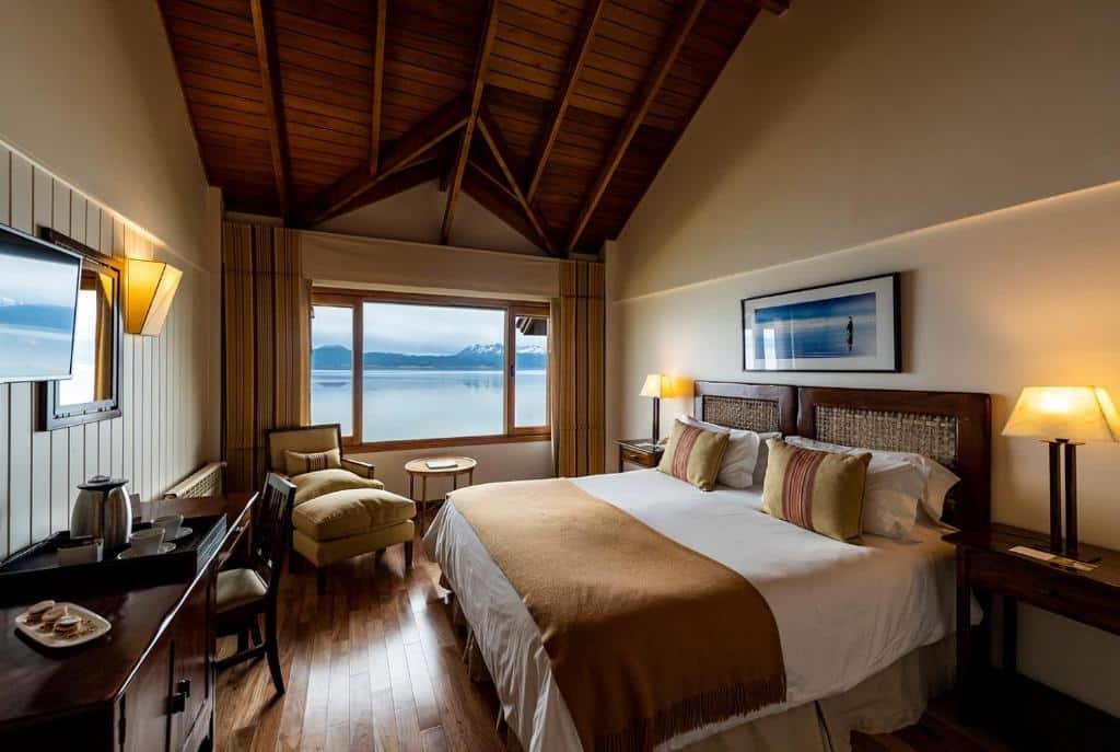 Quarto do Los Cauquenes Resort + Spa + Experiences. Uma cama de casal do lado direito, com um abajur de cada lado, de frente para a cama uma televisão, uma mesa de trabalho com xícaras de café e um prato de doce. No fundo do quarto, uma poltrona, uma mesa e a janela do quarto.