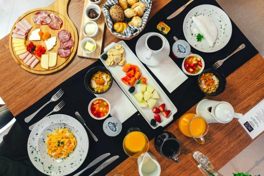 Mesa posta no Lote20 Hotel Boutique, um dos hotéis em Bento Gonçalves, com tábua de frios, guarnições de manteiga, requeijão, geleia e outros, frutas cortadas, salada de frutas, cereais, suco, leite, café, ovos mexidos e tapioca