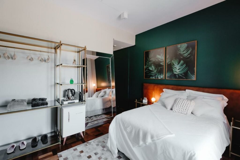 Quarto elegante no Lote20 Hotel Boutique, com uma parede verde escura, quadros com fotos de folhagens combinando, e roupas de cama branca, além de armário aberto, frigobar, chaleira e espelho de corpo inteiro no recinto