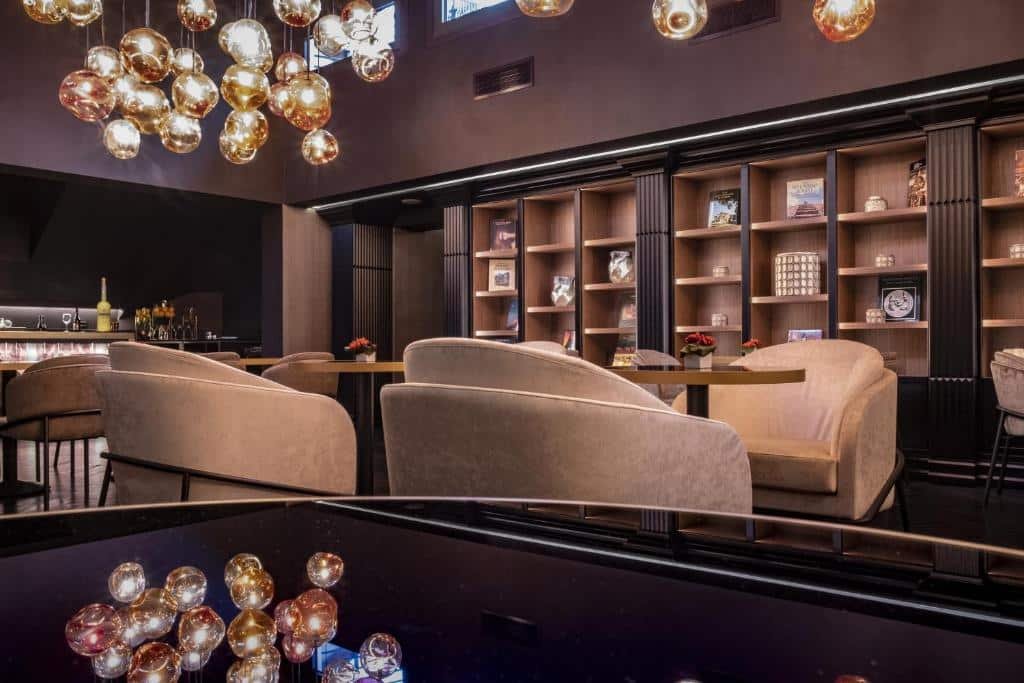 lounge do U-Visionary Roma Hotel com poltronas grandes e cores claras, com mesinhas de madeira e muitas prateleiras e nichos na parede com decorações, os lustres e luminária são redondos e estruturados