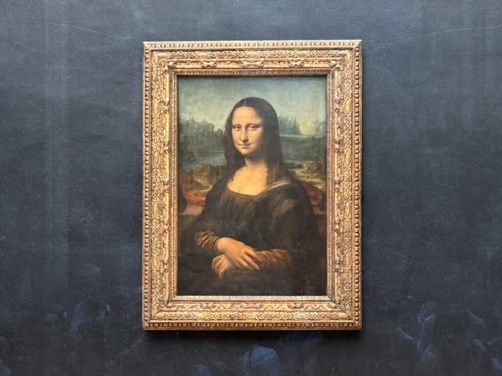 Uma pintura emoldurada da Mona Lisa com um fundo escuro no Museu do Louvre. A obra de arte apresenta uma mulher com um sorriso sutil e mãos postas.