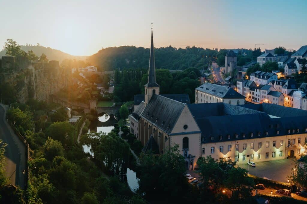 Foto aérea da Cidade de Luxemburgo. Á esquerda, vemos fortes construídos de pedra, e uma rodovia que sobe em sua direção. Na sua frente, há um lago e uma ponte para realizar a travessia, que conecta à cidade. Na direita da foto, vemos construções com arquitetura típica do país. Há uma igreja com uma torre pontuda em destaque, e mais casas e prédios atrás. As luzes das residências e das ruas estão acesas, pois já está quase anoitecendo. O sol se põe atrás das montanhas no fundo da imagem.