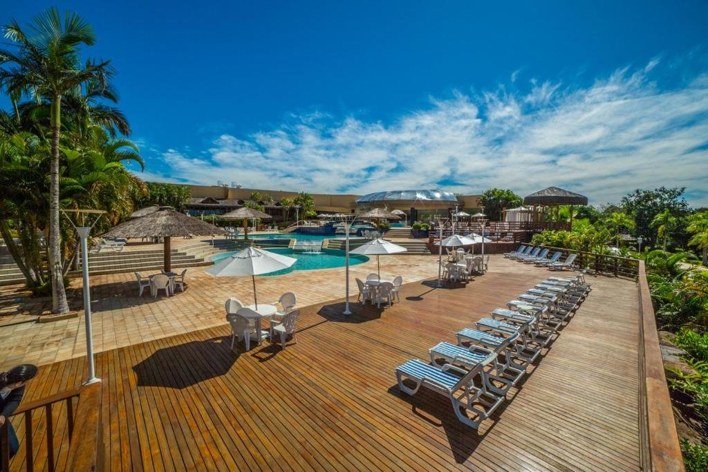 Parte do Mabu Thermas Grand Resort em Foz do Iguaçu que mostra em primeiro plano várias espreguiçadeiras e mesas com guarda-sóis. Ao fundo tem uma piscina ao ar livre e várias palmeiras verdes ao redor do ambiente. Imagem ilustrando post Resorts em Foz do Iguaçu.
