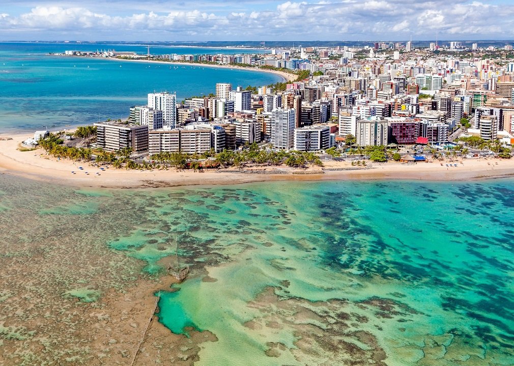 Mar ao redor de uma região costeira de Maceió, que tem diferentes tipos de prédios e casas. O dia está ensolarado e o mar possui águas transparentes, mostrando as barreiras de corais no fundo do mar