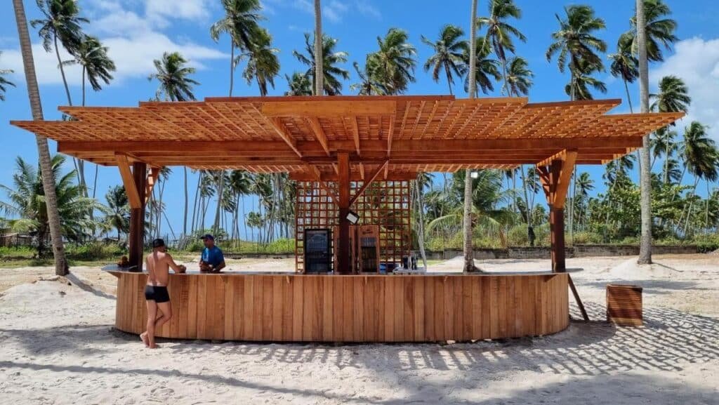 Um bar de praia de madeira no Maceio Mar Resort All Inclusive com duas pessoas em pé perto do balcão, com palmeiras altas e um céu azul claro ao fundo.