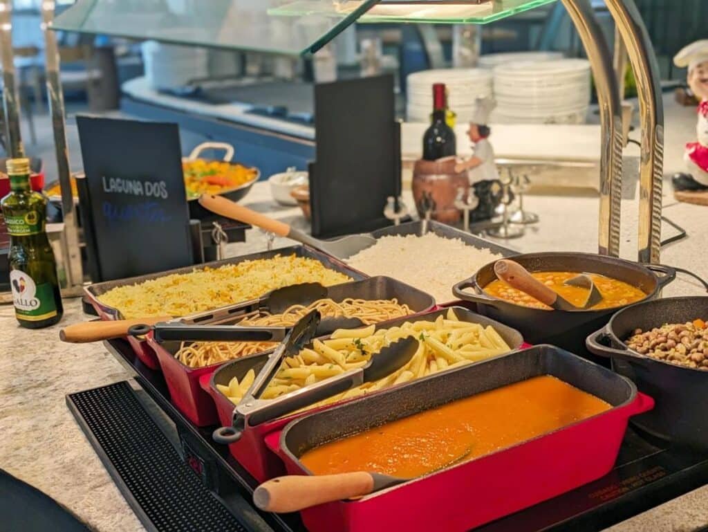 Balcão de bufê no Maceio Mar Resort All Inclusive com bandejas de arroz, espaguete, macarrão penne, sopa, grão-de-bico e condimentos, com decoração de cozinha ao fundo.