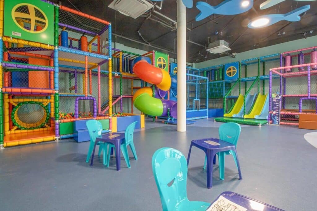 Área de recreação infantil interna no Maceio Mar Resort All Inclusive com estruturas coloridas de escalada, escorregadores e mesas com cadeiras. Piso azul e decorações suspensas.