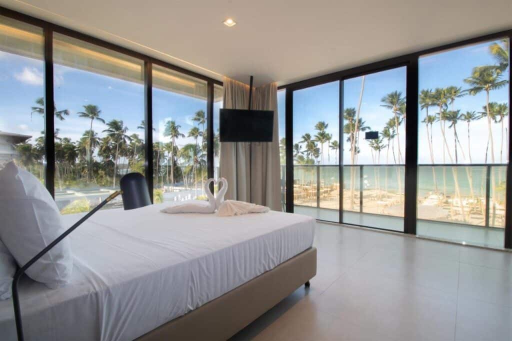 Quarto moderno no Maceio Mar Resort All Inclusive com uma cama grande, toalhas em formato de cisne, paredes de vidro com vista para o mar e as palmeiras e uma TV montada na parede.